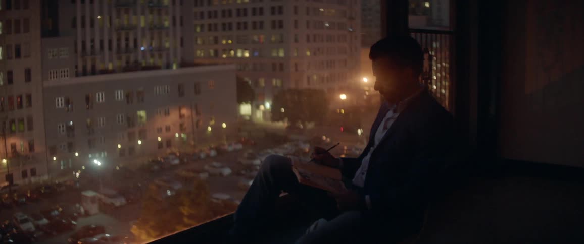 A man writes in a notebook while overlooking a city from a large window