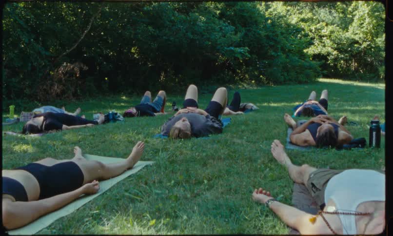 A group of people lie on yoga mats in a grassy clearing surrounded by trees resting on their backs as sunlight filters through the foliage