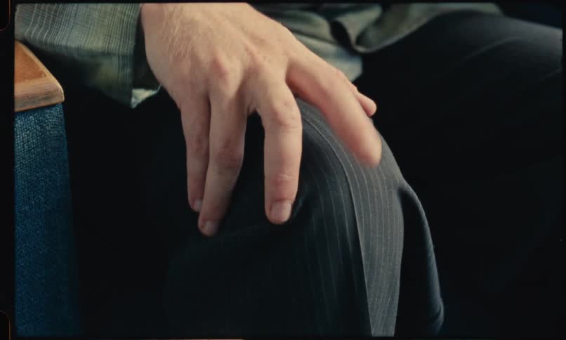 A closeup shot shows a persons hand resting on their lap while their fingers rhythmically tap against their pinstriped trousers the repetitive motion and vintage film aesthetic suggest a sense of nervous anticipation or impatience