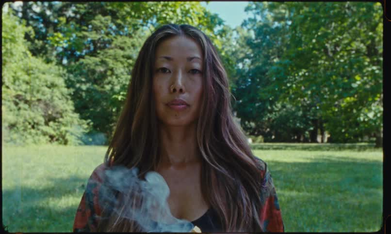 Woman blowing smoke from a lit object while holding a container in a grassy area with trees in the background