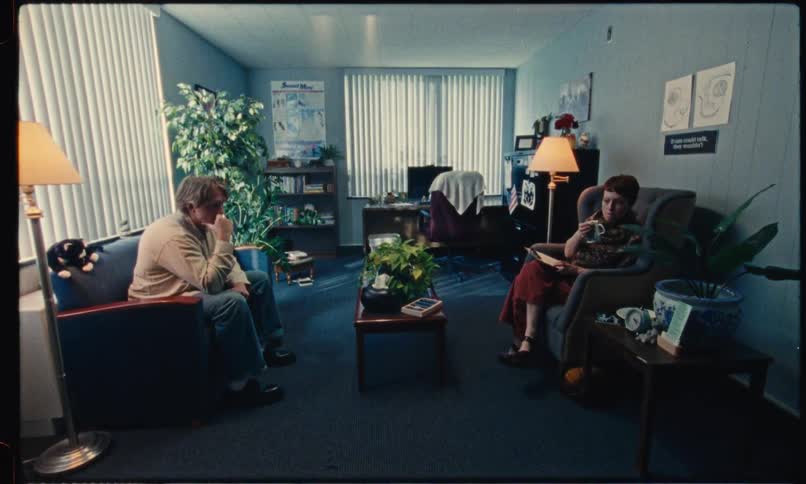 A man sits on a couch across from a woman with a clipboard