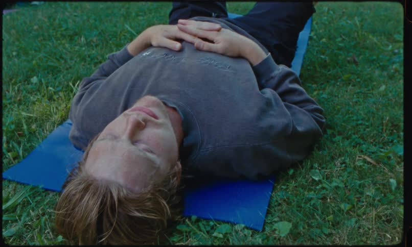 A blonde man lies on a blue yoga mat in a grass field with his eyes closed and hands resting on his stomach while other people practice yoga in the background during an outdoor session
