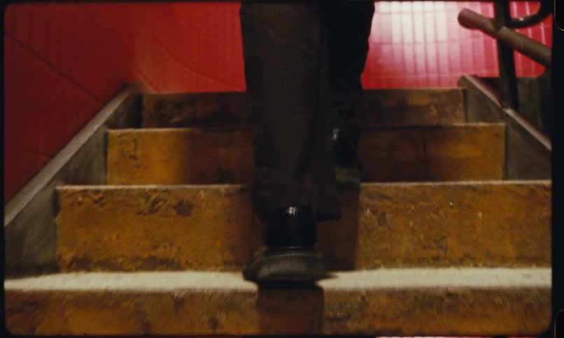 A view from behind showing the legs and feet of a person in dark trousers and black shoes as they walk up a flight of worn yellow stairs in a building with red tiled walls