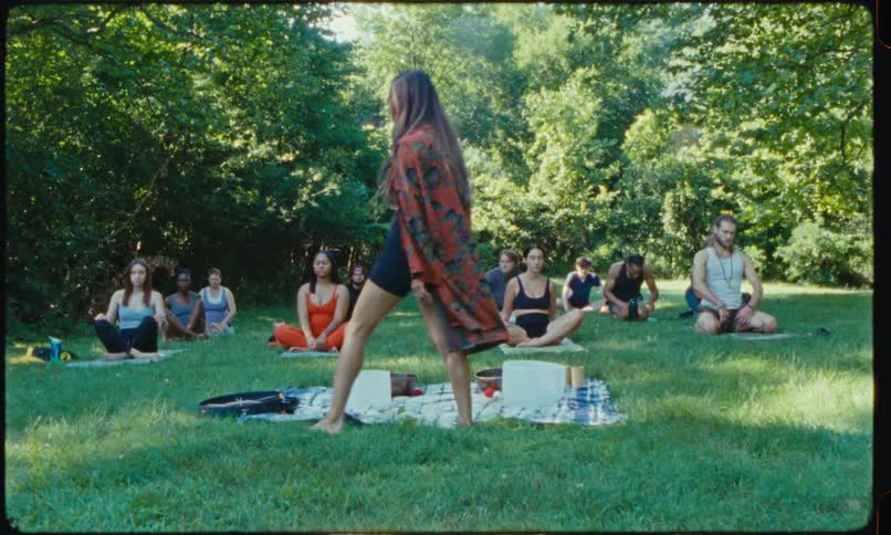 A woman wearing a long patterned robe stands before a group of people sitting on yoga mats in a grassy park clearing surrounded by lush green trees during a bright day