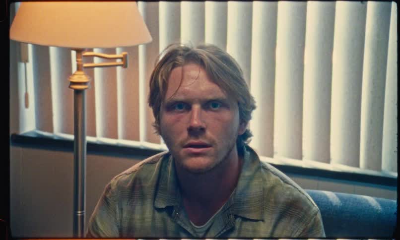 A man with blonde hair and blue eyes sitting in a room with a lamp and vertical blinds the camera slowly zooms in on his face as he stares directly forward with a somber or shocked expression