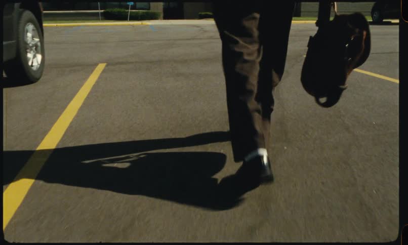A person exits a car and walks across a parking lot while carrying a bag