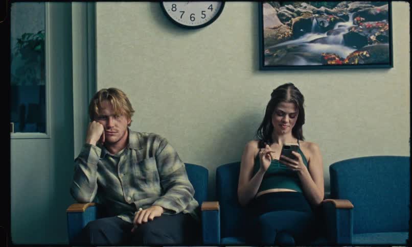 Man and woman seated in waiting room chairs he looking down and she talking on the phone beneath a clock and waterfall picture on the wall