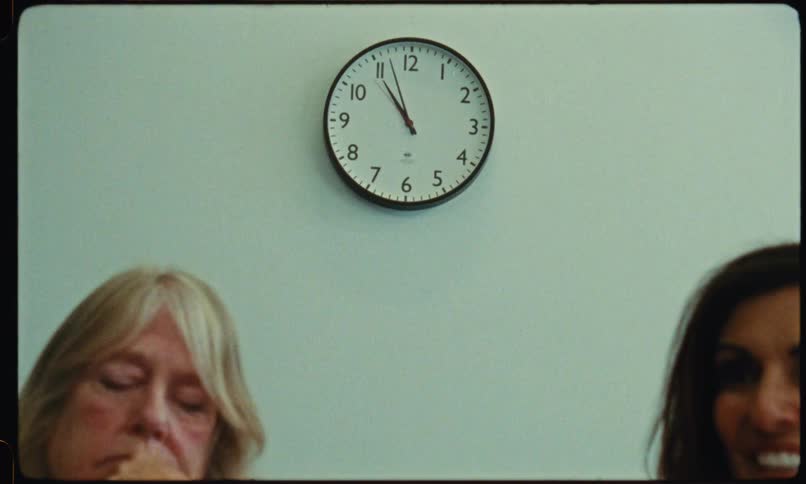 Two middleaged women sit behind a desk inside an office conducting an interview with an analog wall clock on the wall behind them
