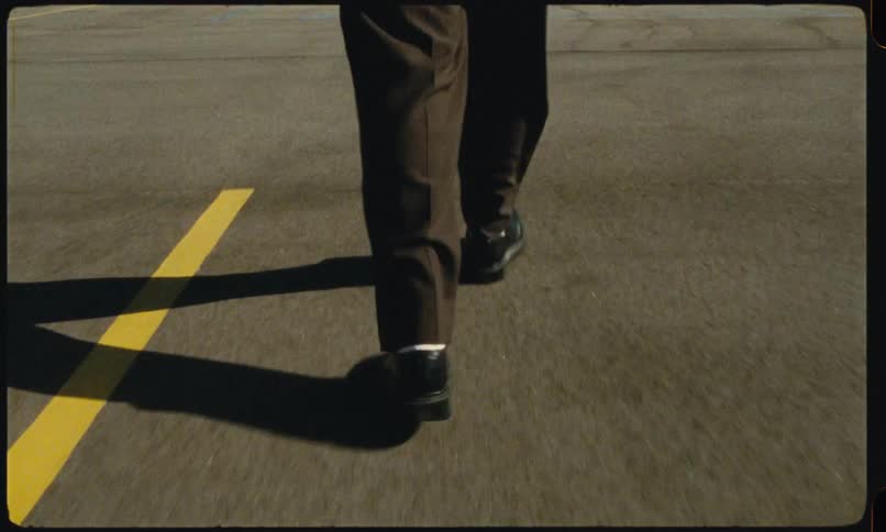 A view from behind shows the legs and feet of a person wearing brown trousers and black shoes as they step out of a car and walk across a parking lot on a sunny day