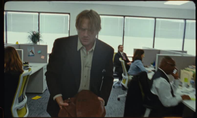 Man in a suit discarding an object into a trash can inside an office with coworkers in the background