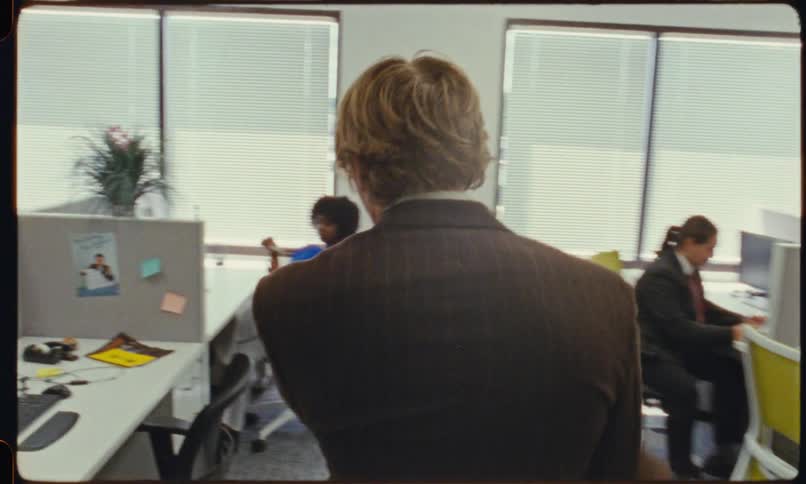 A man in a brown suit coat walks through an office to sit at his cubicle