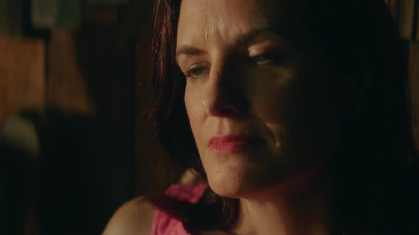 A woman smiles and stares while inside a home with a man in the background drinking a beverage
