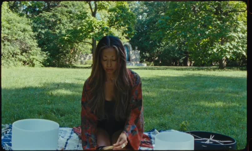A woman sits crosslegged on a blanket in a sunlit park surrounded by singing bowls and sound healing instruments as she gently plays one