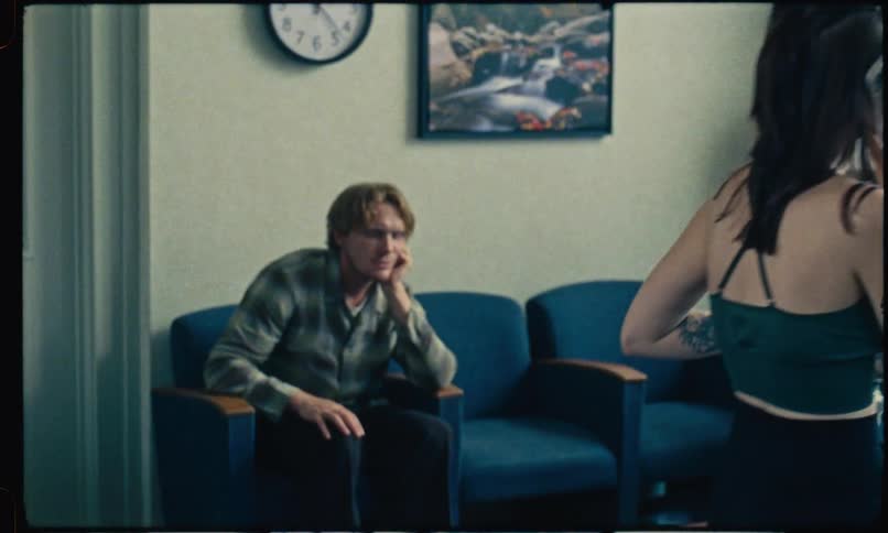 A woman who had been speaking through a glass partition now stands in a waiting room talking on her phone while holding a drink as a man beside her sits slouched in a blue chair beneath a wall clock