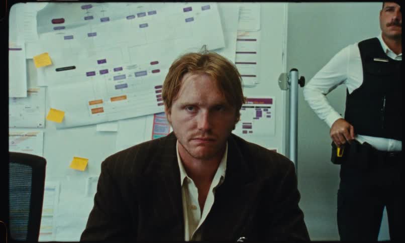 Man seated at a table is held by the arm by another man in a room with papers pinned on the wall