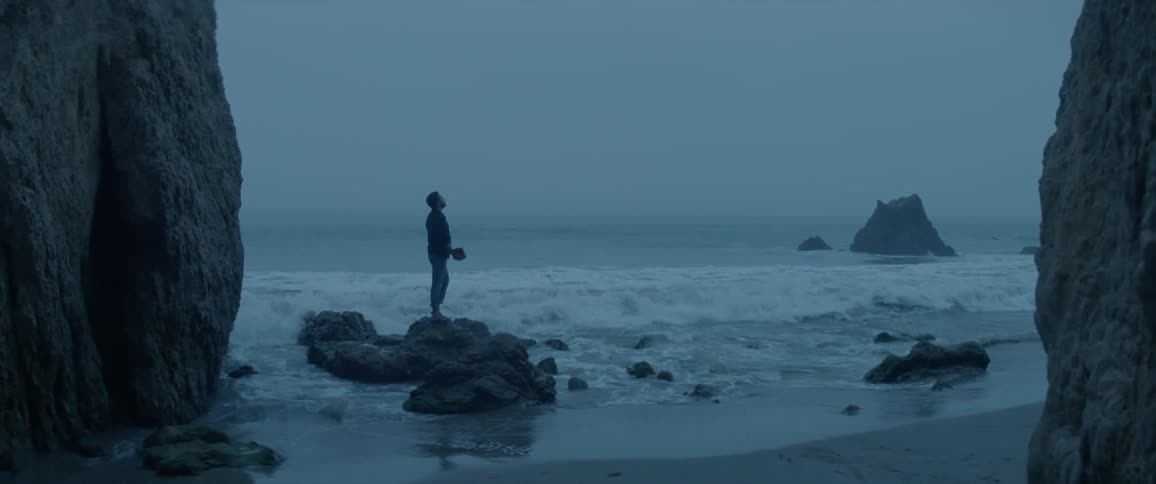 A man stands atop rocks as waves crash ashore