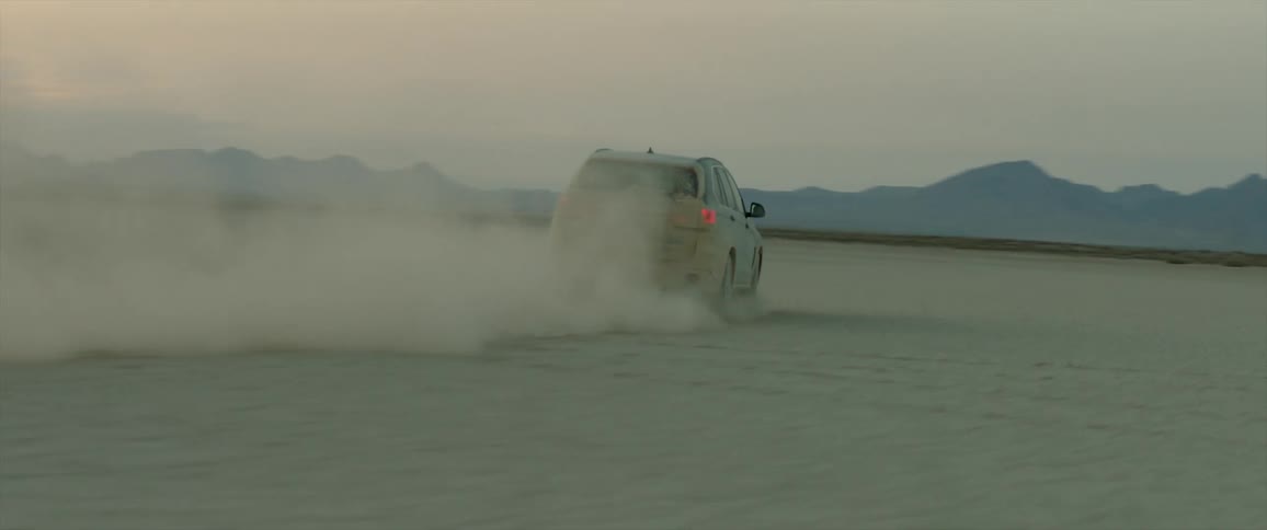 A car drives through a large dusty expanse surrounded by mountains at sunset