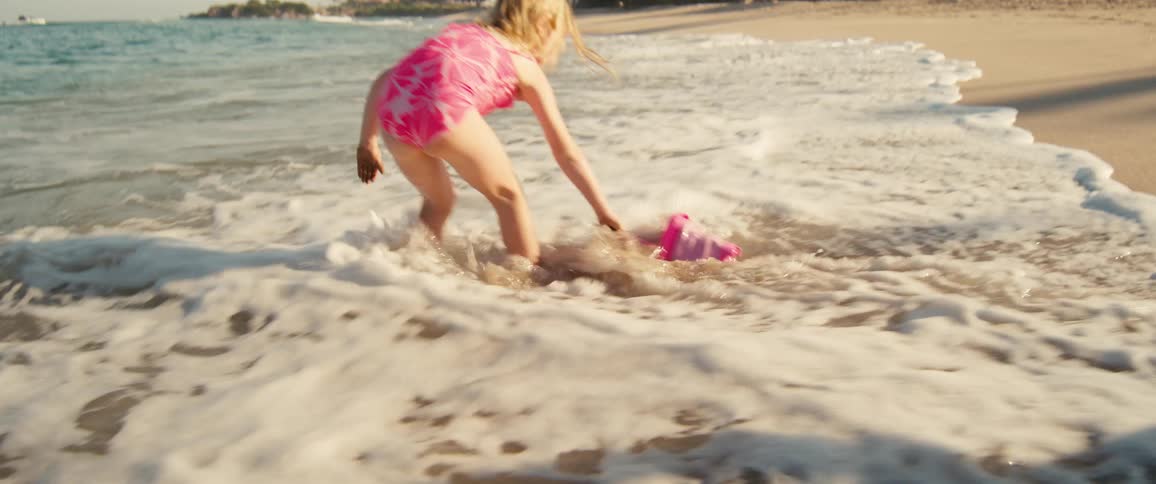 Girl in a pink swimsuit crouching at the shoreline holding a bucket in the water