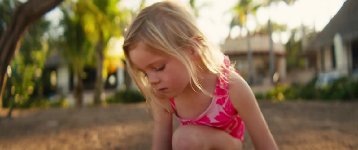 A young girl in a pink swimsuit crouches in the sand carefully shaping it with her hands as palm trees and tropical buildings blur in the background
