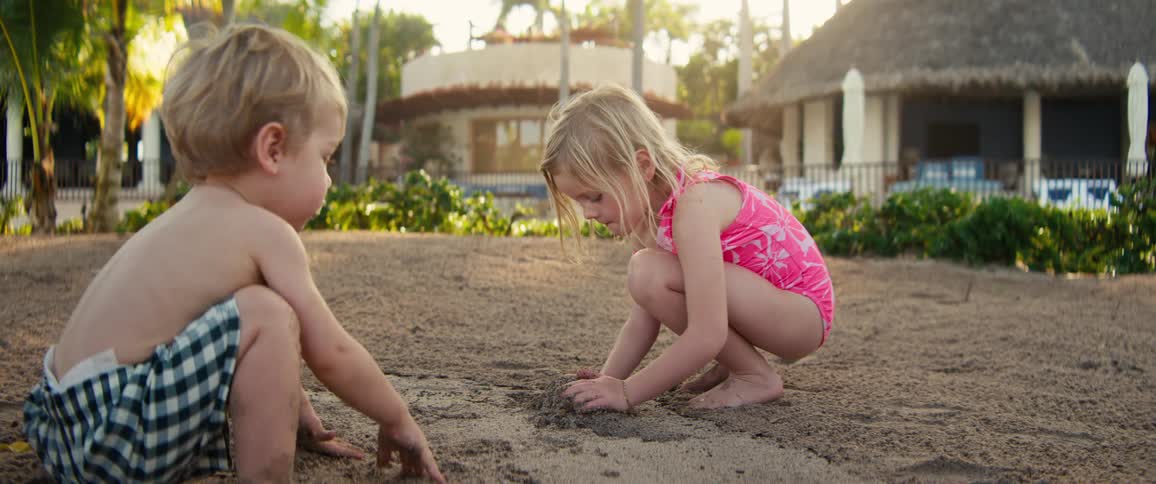 Two young children crouch in the sand near a tropical resort concentrating as they dig and shape the ground with their hands