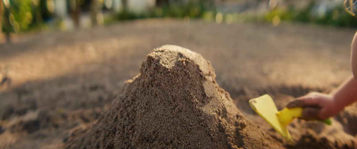 A mound of damp sand sits in the center of the frame showing texture and small variations in colora child uses a bright yellow toy shovel to pat and shape the sand pile eventually moving around it as they play in the sunlight