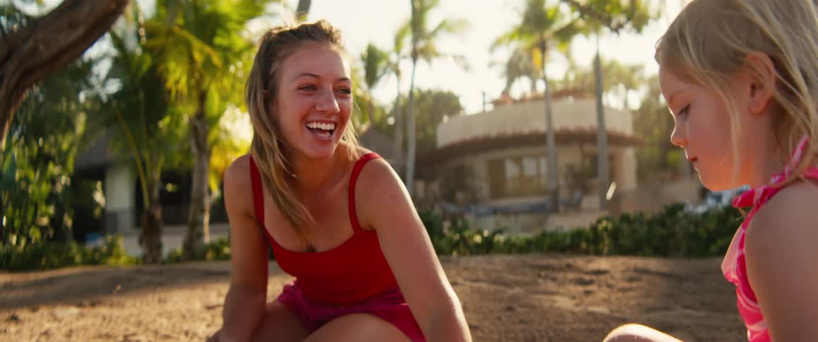 A woman wearing a red tank top and a young girl play together in the sand on a tropical beach with palm trees and a building in the background during a bright sunny day