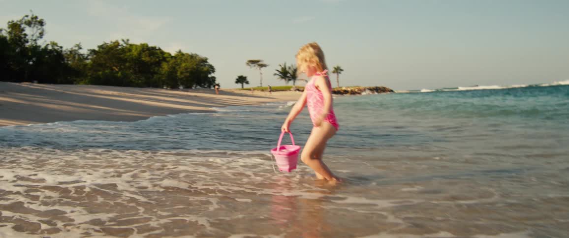 Girl in a pink swimsuit holding a bucket in the shallow water at the beach