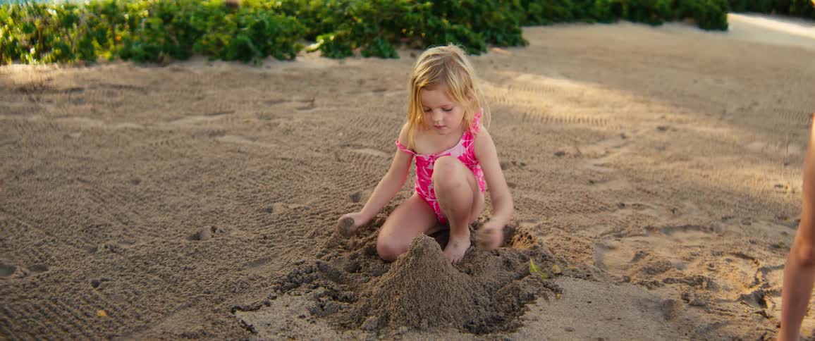 Little girl playing in the sand building a mound as another girl stands nearby