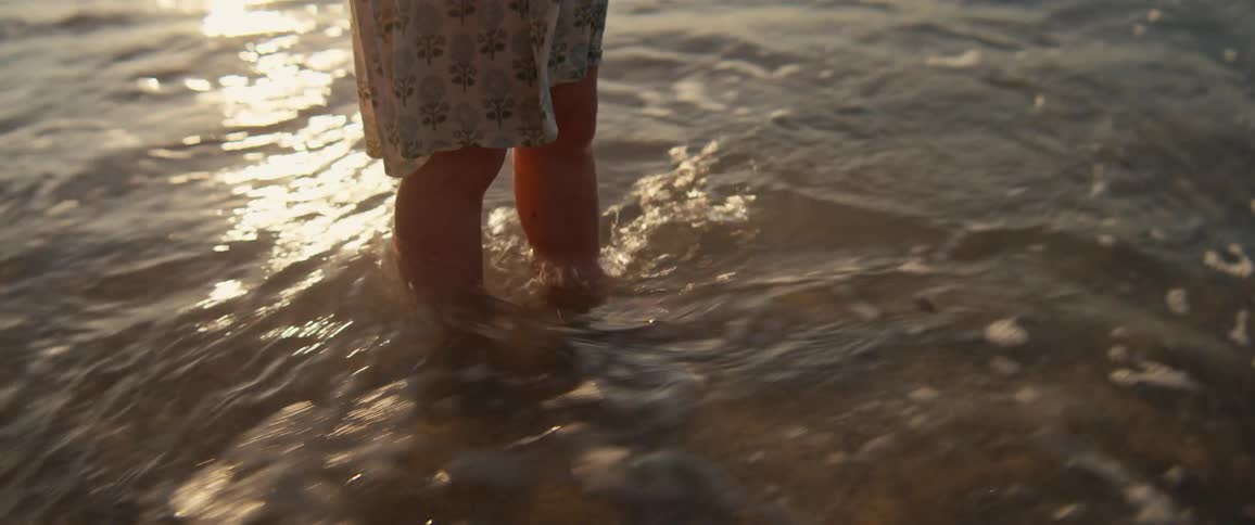 A person wearing shorts stands on the beach at sunset as waves wash over their feet and recede back into the ocean