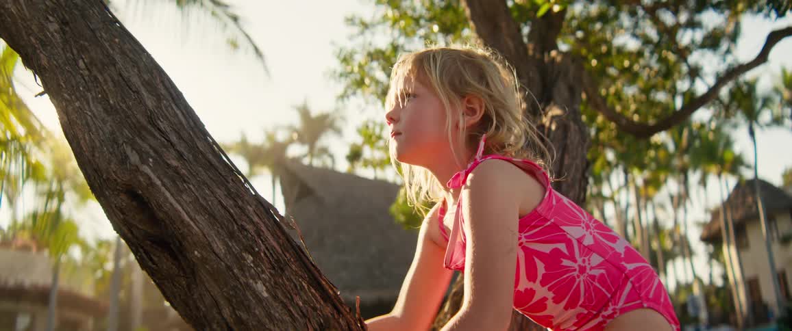 Small girl in pink swimsuit climbs a tree in a tropical neighborhood