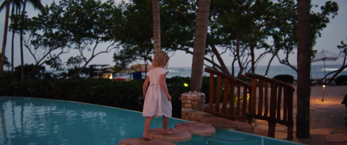 Young blonde girl crosses over a swimming pool located near the beach on a path in the evening