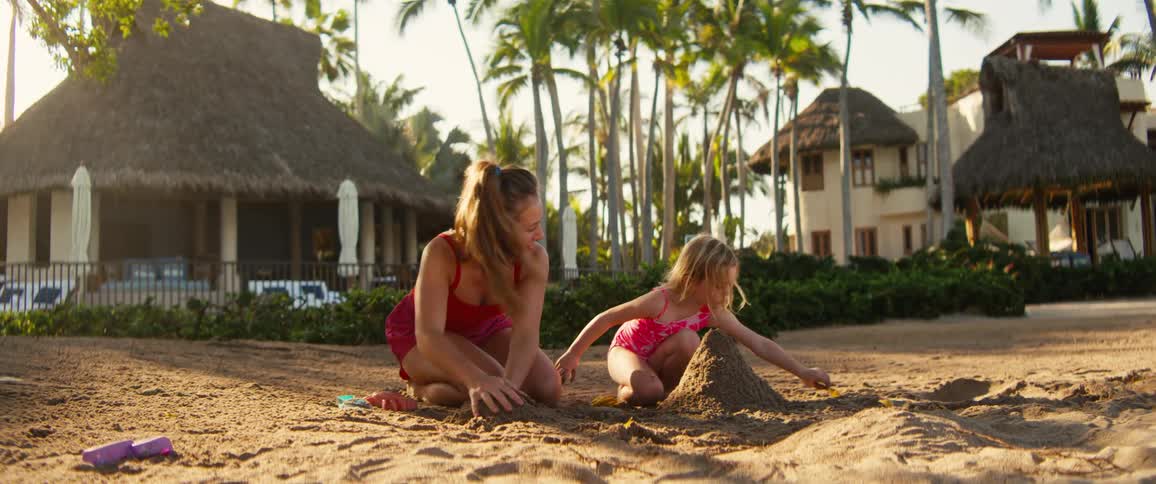 A woman and a young girl in pink swimwear are kneeling on a sandy beach focused on building a sandcastle togetheras the sun sets behind palm trees and beach huts the girl stands up and runs offscreen leaving the woman to continue molding the sand