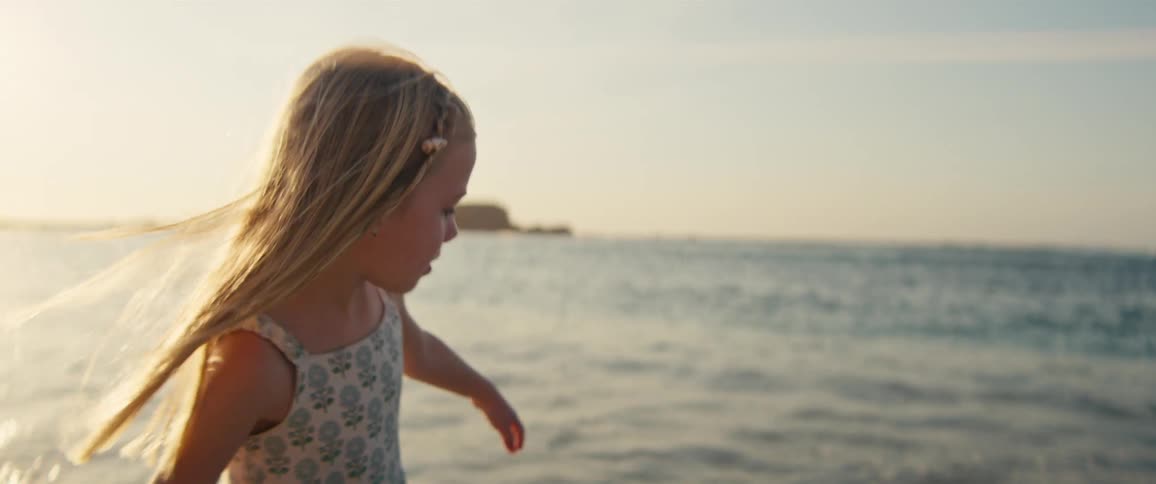 A young girl with long blonde hair wearing a floral dress walks along the shore as ocean waves gently lap at her feet during a bright golden sunset with a small island visible in the distant sea