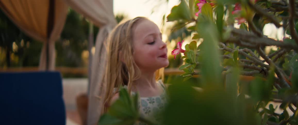 A young girl with long blonde hair leans in to smell a vibrant pink flower on a desert plant during a golden sunsetshe smiles playfully after catching the scent bathed in warm light and a soft lens flare