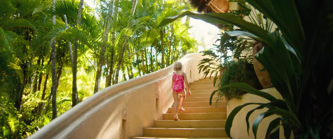 Girl in a pink swimsuit climbing stairs outdoors surrounded by tropical vegetation