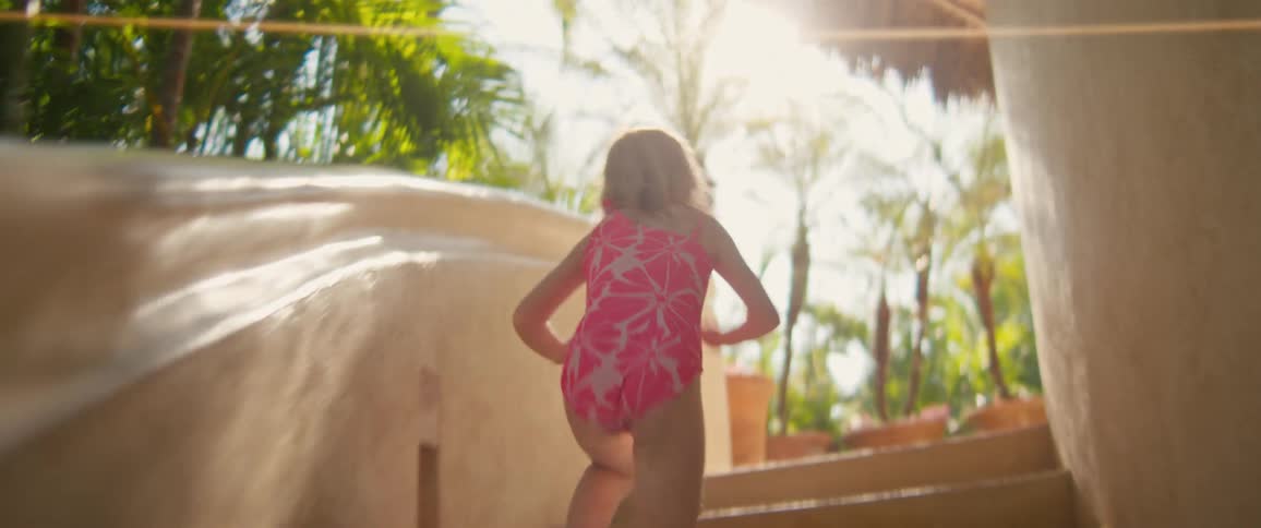 Small blonde girl wearing a pink swimsuit walking and then running upstairs to a balcony where she walks and then runs again outdoors during day time