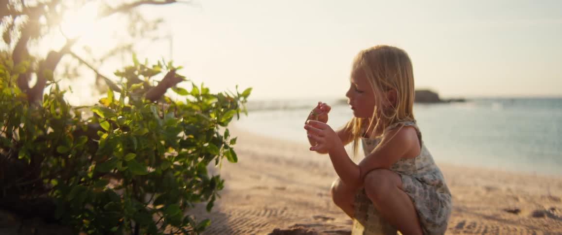 Blonde girl crouches on the beach while picking leaves from a plant on a sunny summer day