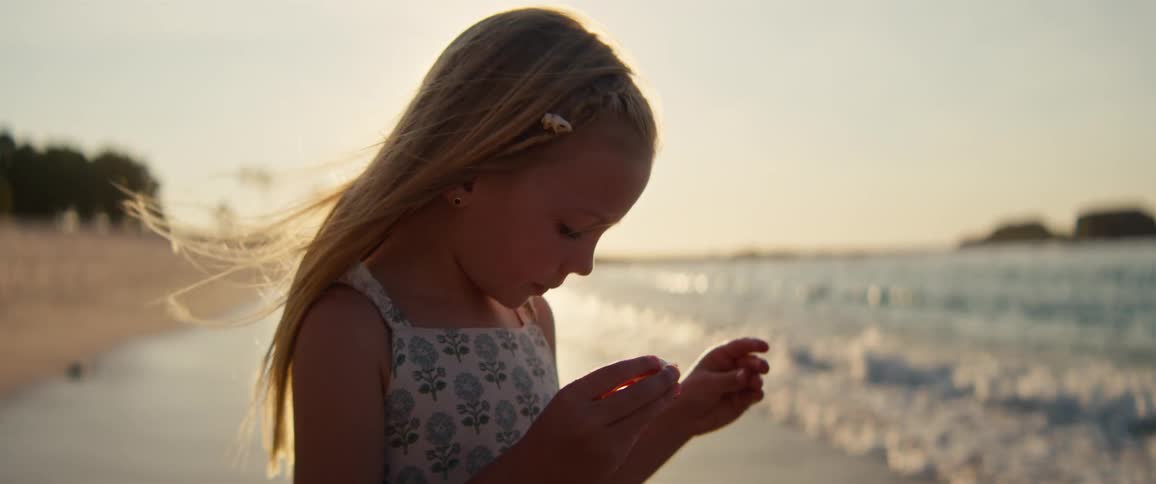 Blonde girl stands on the beach on a summer day examining a seashell