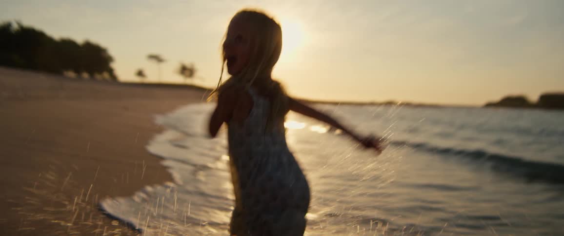 A young girl wearing a floral dress runs barefoot along a sandy beach at sunset while splashing in the shallow ocean waves during a golden hour evening