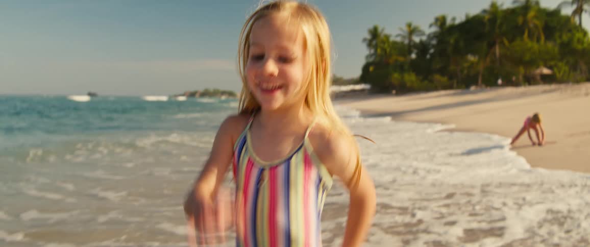 A young girl wearing a colorful striped swimsuit smiles and runs through shallow ocean waves on a sunny beach with tropical palm trees in the background