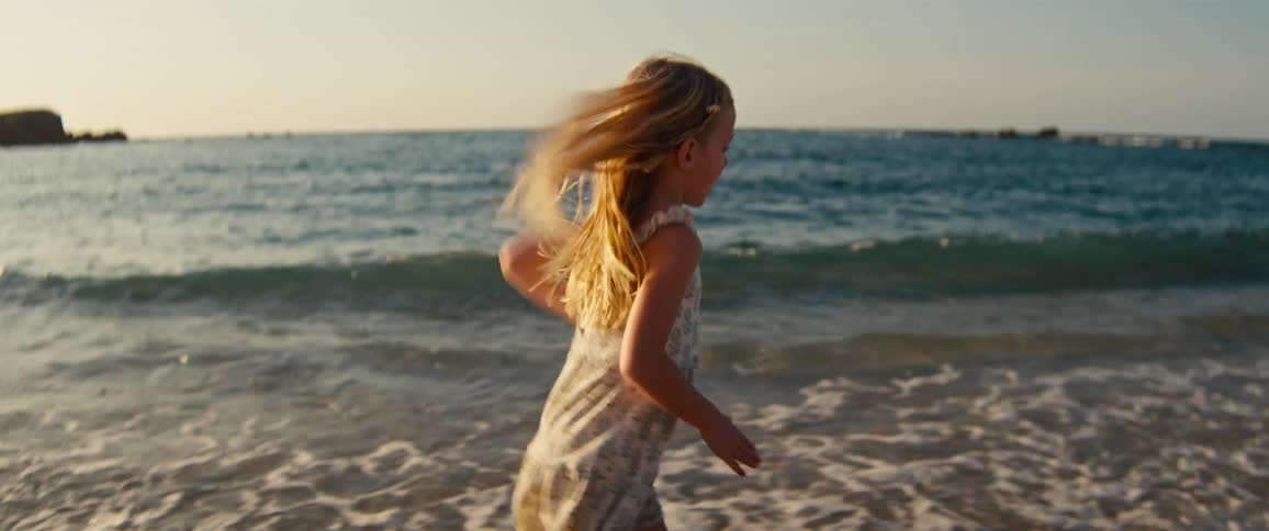 A blonde girl in a floral dress runs along the shoreline during golden hour as waves crash on the sand with a rocky coastline and blue ocean in the background
