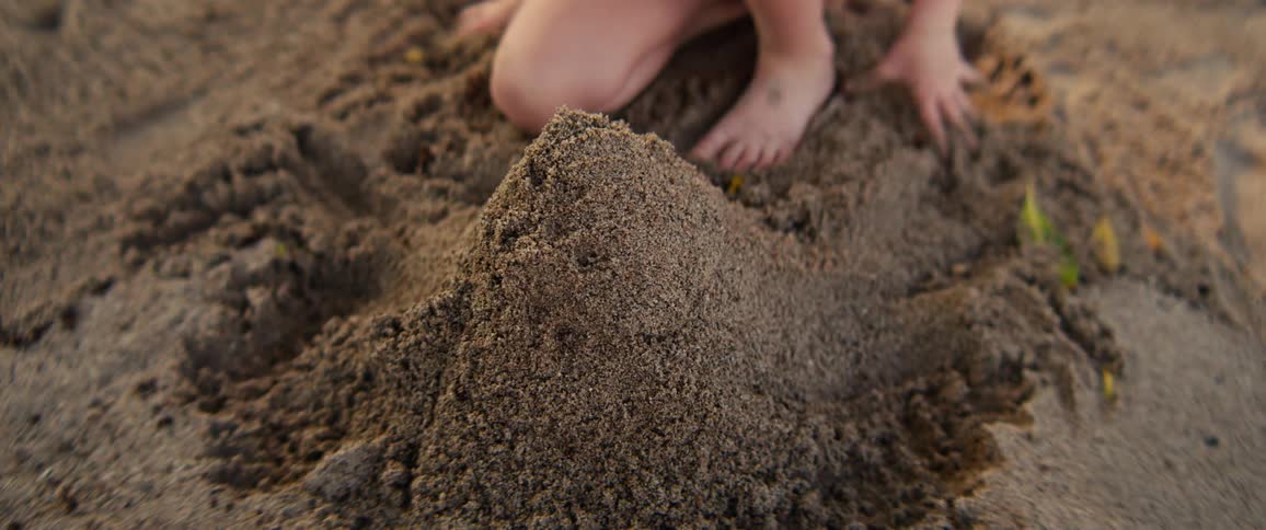 A young girl uses her hands to shape a pile of dark wet sand into a small mountain while sitting on a beach
