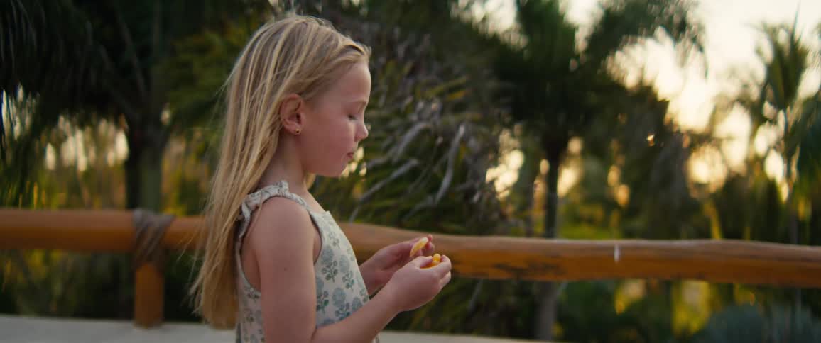 Blonde girl holding a small object in her hands near a wooden railing with palm trees in the background