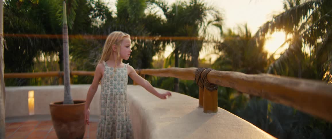 Young blonde child wearing a dress standing on a balcony looking at a view of palm trees and the sunset outdoors