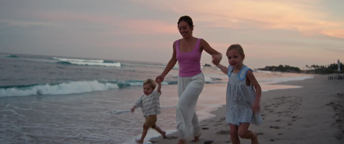 A woman walks along the beach at sunset holding hands with a young child while another little girl runs happily beside them near the shoreline
