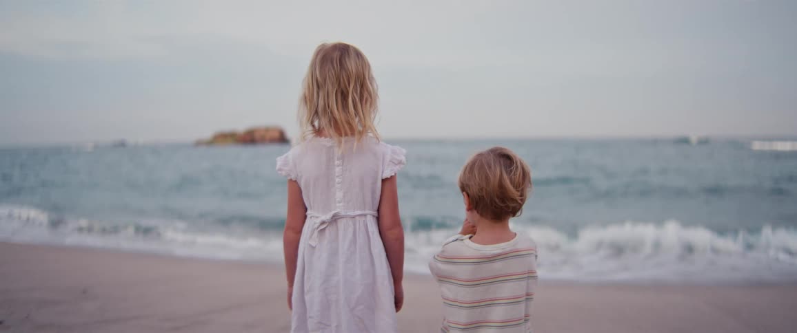 Two young children stand side by side on the beach gazing out at the ocean waves and a distant rocky island