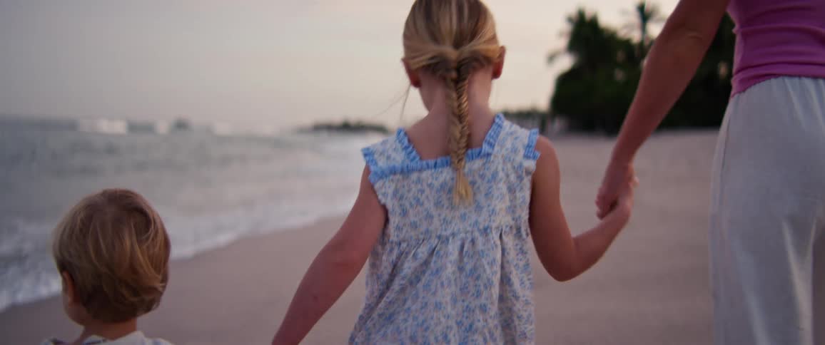 A young girl with braided hair wears a blue floral dress while walking on a beach at dusk holding hands with a small boy and a woman as waves lap against the shore
