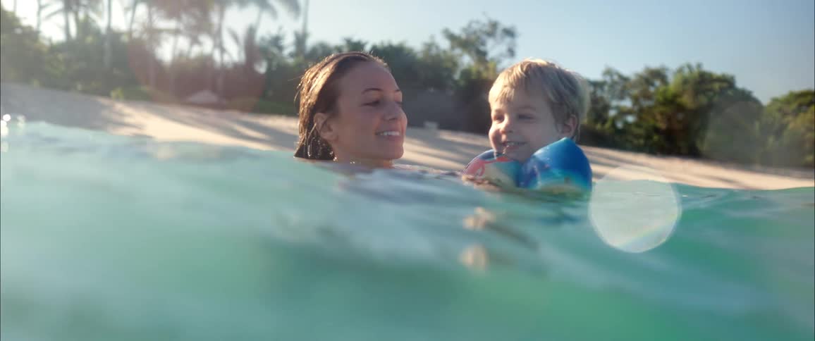 Young adult woman bathing on the sea and holding in her arms a toddler with swimming wings near the beach outdoors during day time