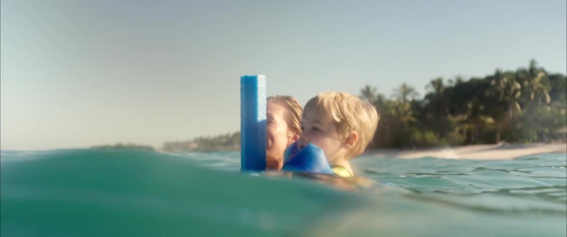An adult and a young child smile while floating in the ocean holding onto a blue pool noodle as gentle waves ripple around them
