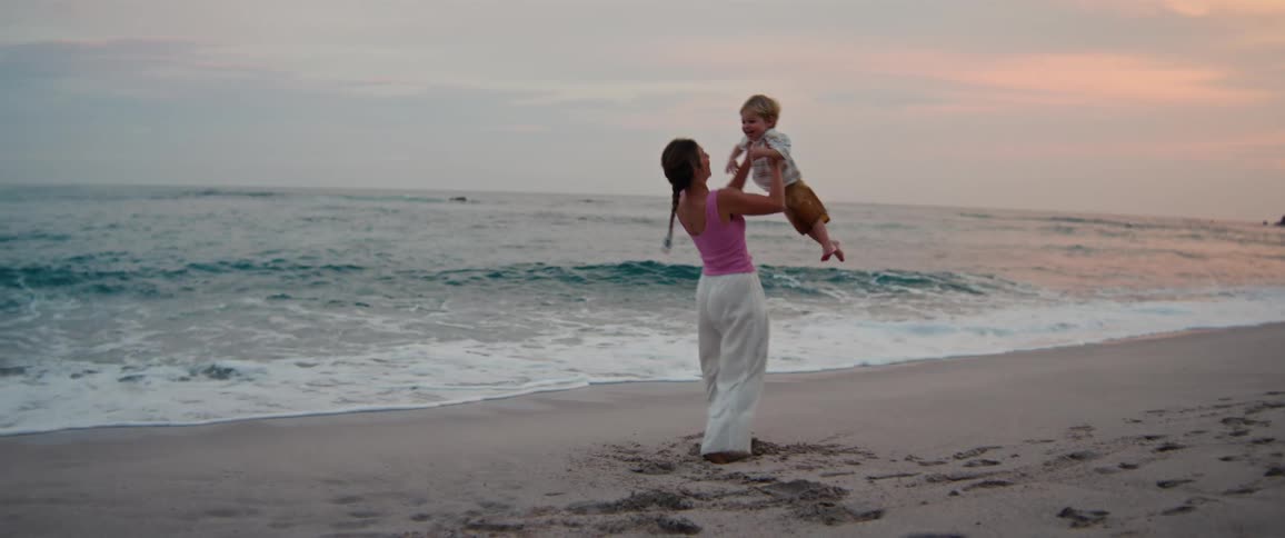 Woman stands on the beach holding her young son in the air spinning around